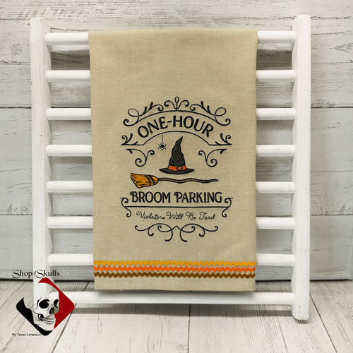 Beige kitchen towel embroidered with a witch hat and broomstick over the words: one hour broom parking.